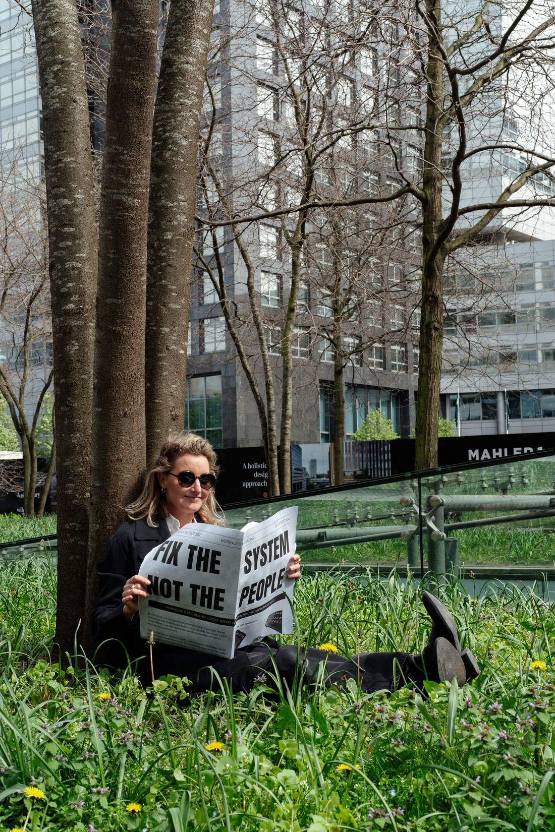 Dian zit in het gras onder bomen voor kantoorgebouwen en leest een krant met de kop Fix the system, not the people